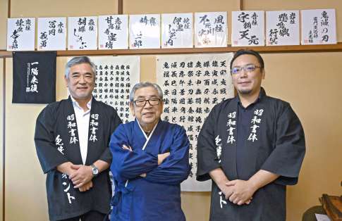 綱紀栄泉さんを中央に笑顔を見せる会長の坂口茂樹さん（左）と社長の太樹さん。壁の上段に飾ってあるのは「鬼滅の刃」で使われたフォント＝さつま町虎居町