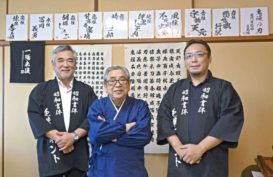 綱紀栄泉さんを中央に笑顔を見せる会長の坂口茂樹さん（左）と社長の太樹さん。壁の上段に飾ってあるのは「鬼滅の刃」で使われたフォント＝さつま町虎居町