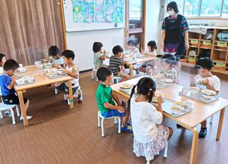 感染防止のアクリル板が設置されたテーブルで給食を食べるこども園の子どもたち。コロナ禍で現場の負担は大きくなっている＝南さつま市