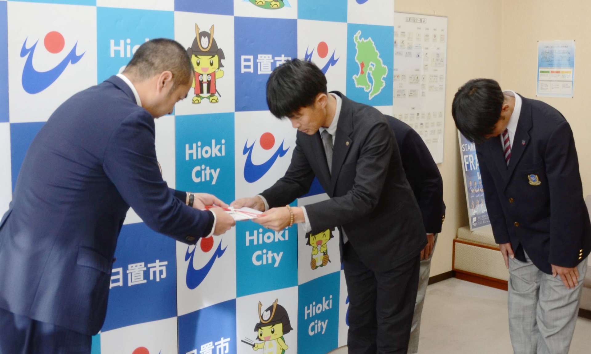 鹿城西高サッカー部の新田祐輔監督（中央）に補助金１００万円の目録を手渡す永山由高・日置市長＝市役所