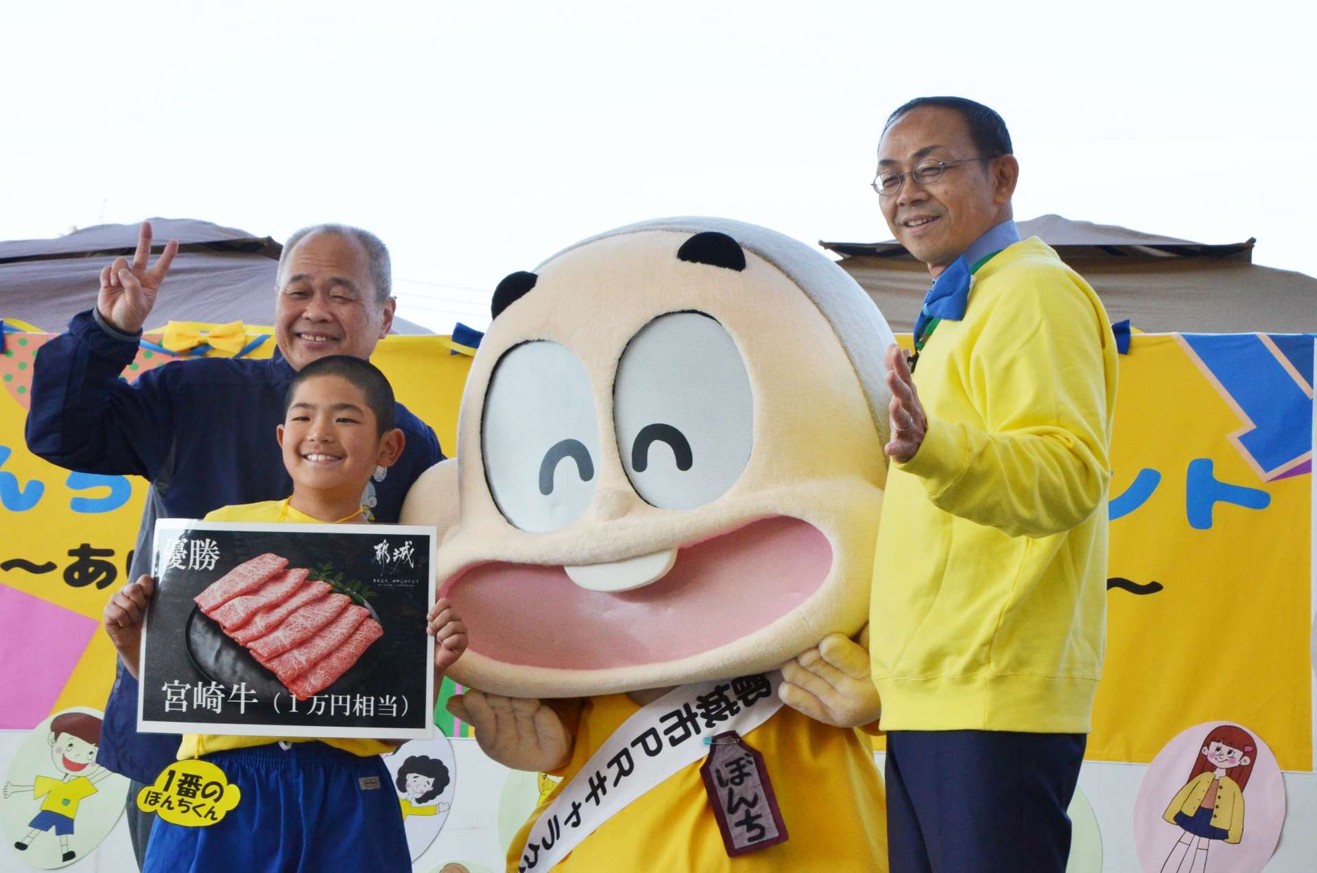 ぼんちくん作者の今村幸一さん（左後方）と池田宜永市長（右）に祝福されるそっくりさんコンテスト１位の牛谷将馬さん（左手前）＝都城市都北町