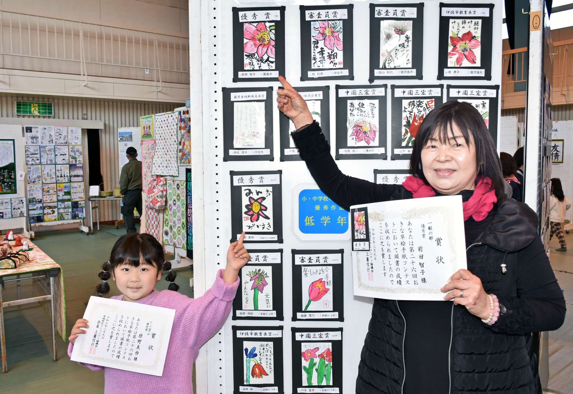 〈関連〉優秀賞を受賞した作品をそれぞれ指さす前田智子さん（右）と孫の原野美怜ちゃん＝伊佐市の本城小学校体育館