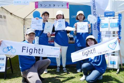 ３小学校の児童が考えたノベルティーグッズをＰＲするランスタッドの社員＝２７日、鹿児島市のウオーターフロントパーク