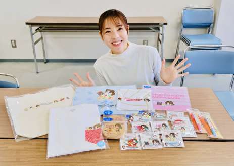 販売中のコラボグッズを広げて笑顔を見せる迫田さおりさん（スポーツビズ提供）