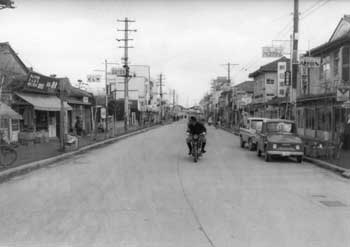 昭和37年、鹿児島市と合併をひかえる谷山市のメーンストリート。大鹿児島市建設構想のもと、道路拡張や舗装が着々と進む。