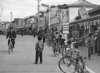 昭和37年、旧吹上町の伊作商店街。「ブロックやモルタル塗りの近代建築がならび、コンクリートの舗装が完成、町外買い出し流出を防ごうと、専門店会の発足など、堅実な経営がすすんでいる」と紙面で取り上げている。