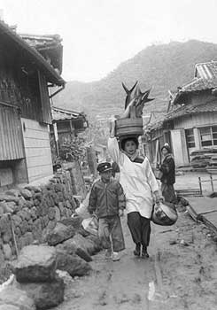 昭和37年、旧坊津町。「坊津町は県下でも有数の急傾地帯。このため頭の上にのせて物を運ぶ“かんめ”という風習が続いている。段々畑でタキギを運んだり揚げ場からでっかい魚をおけにのせて町を行く風景はちょっと他の地方ではみられない」と紹介。