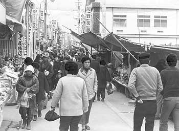昭和45年12月末、旧加世田市本通り。「加世田市名物の“としの市”は、南薩各地から買い物客およそ二万人がつめかけ身動きできぬ混雑ぶり。農村景気の好調と出かせぎ者からの送金でホクホクの買い物客はサイフのひももゆるみっ放し」と取り上げている。