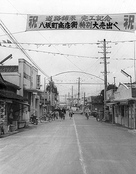 昭和35年、大口市の八坂通り。紙面では「道路舗装工事がこのほど完工。大口市の目抜き通りにあたり、商工業者の喜びは大きい。延長百四十三メートル。これで同市の中心街の舗装は八分通り完成をみた」と報じている。