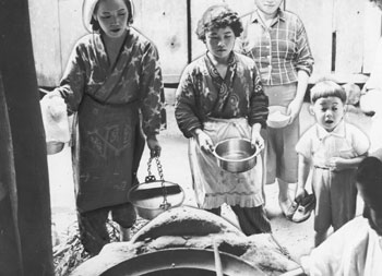 昭和３０年６月、旧志布志町宮地集落の共同炊事。紙面によると「同集落は田植え時期を迎え、共同炊事と託児所を設け能率をあげている。初めての試みで、十四戸七十人が参加した」と伝えている。