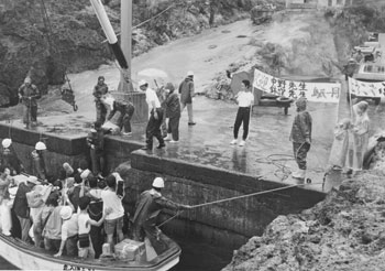 平成元年、のぼりを掲げて先生を出迎える小宝島の人たち。「昭和五十四年に廃校になって十年ぶりに宝島中小宝島分校が再興。鹿児島市から生徒が１人転入し、若い先生が２人赴任してきた」と紙面は報じている。