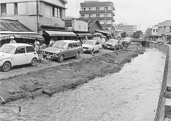 昭和４８年６月、名瀬市中心街の渋滞は深刻で、駐車場にできないかと目をつけられている永田川。当時の紙面では「市中心の商店街で一番困っているのは青空駐車。観光客もびっくりの自動車の多さで、前年の乗用車伸び率は３７％で全国の伸び率を大きく上回った。一帯は狭い土地に家屋が建て込んでおり、混雑解消と駐車場確保のために頭を痛めている」などと報じている。