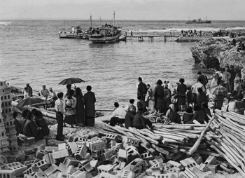 昭和３０年３月１２日、定期船が接岸できず、ハシケで荷下ろしや乗・下船をする喜界島の湾港。当時の紙面では「鹿児島と結ぶ船の汽笛が響くと、港は時ならぬにぎわいを見せる。サンゴ礁の沖合にイカリを降ろした船まで、客も荷物もハシケで送り迎えられる。岸壁には建築資材が雑然と積まれ、港は黒糖やトマトの出荷にいそがしい。出稼ぎの若者を見送る家族の姿が印象に残る出船風景」と紹介していた。