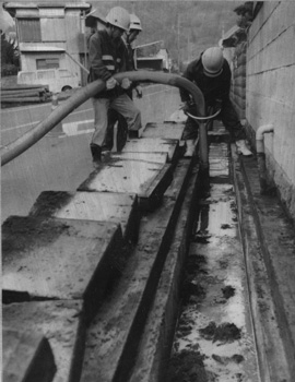 昭和６０年２月、垂水市では桜島降灰が続き、側溝に灰がたっぷり詰まり、取り除く作業に追われる。紙面では「記録破りの噴火で、垂水市には１月だけで前年１年分の２．７倍にも達する降灰。１平方メートル当たり約３４キロ。過去７年間の降灰量を上回り、観測史上最多の“豪灰”だ」と伝えていた。