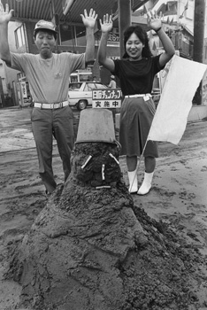 昭和６０年７月３０日、ドカ灰の直撃を受けた鹿児島市城南町のガソリンスタンドの店頭に登場した“灰ダルマ”。当時の紙面は「『手も足も出ません』と白旗を掲げるこの灰ダルマ。かきあつめた灰の量１５０キロを見て、所長らがちゃめっ気を出して仕立て上げた。通りがかりの車からのぞきこむ人もいる」と紹介している。