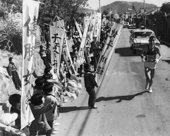昭和４３年２月１８日、「がんばれ大島」「健闘川辺」など全チームのノボリをつくって声援する開聞小の児童たち。紙面では「県下一周駅伝は絶好の“駅伝日より”で開幕した。初日のハイライトは最終区、知覧署ー加世田間１７．１キロ、長距離界のエース船迫、浜田、田中の３選手が郷土の期待をになって出場。根性の見せ場を展開した」と紹介していた。