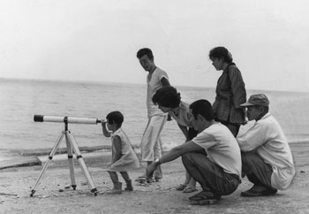 昭和４４年９月、かいもん丸の入港を見物する喜入町（現鹿児島市）民。「入港の模様が見渡せる国道沿いの護岸では、付近の人や通りがかりのドライバーが目前に大きさを増すタンカーにしばし感嘆。子どもや主婦をまじえて海岸に望遠鏡を据え、ムシロを敷いての見物」と紙面は報じている。