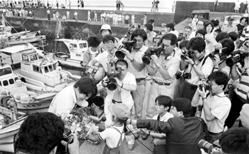 昭和６３年６月１１日、ヨット仲間、取材陣、幼稚園児と多くの人に励まされる今給黎さん。紙面では「世界初の女性単独太平洋往復横断。薩摩おごじょ今給黎さんのボッケな挑戦に、見送りに駆けつけた市民たちも興奮気味。小柄な体で、大きな夢をかなえようとして大海原へ羽ばたく姿に、大きく温かな励ましの声をかけた。岸壁はテレビ各社、雑誌関係のカメラマンもぎっしり。上空には取材ヘリも２機舞い、高まる気分をかきたてた」と報じている。