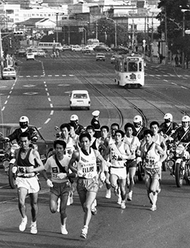 昭和４９年２月１０日、鹿児島市武之橋で谷山中継点をめざし鹿児島市内を一団となって走る選手。紙面では「初日は、午前８時半、鹿児島市易居町の本社前を出発、南薩路を駆けて加世田入りした。春を告げる駅伝は、石油危機のあおりで２日間に短縮されたが、沿道の声援は例年以上に熱烈。力走する選手に向けて拍手が絶えなかった。」と報じている。