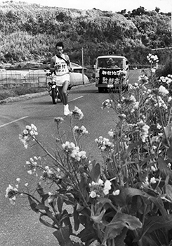 昭和４９年２月１０日、駅伝第１日、開聞町上野で早春の菜の花を横に力走する肝付の高根選手。紙面では「早春の南薩路を駅伝が駆けた。ベテランも新人も、郷土の期待を背に精いっぱいの力走を続ける。錦江湾を左手に見て、花の咲きかおる指宿を抜け枕崎から加世田まで。沿道は、春の訪れを告げる駅伝に沸いた」と報じている。