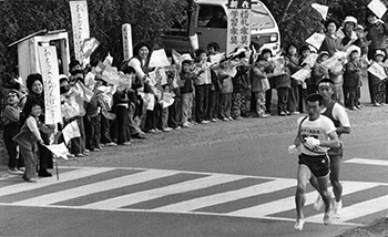 昭和５０年２月１５日、頴娃町御領で家族の声援を受けて力走する指宿・浜崎選手。第２２回鹿児島県下一周駅伝第１日は、絶好の駅伝日和に恵まれ、鹿児島市の本社前をスタート。早春の日ざしを浴び、錦江湾沿いに南下、菜の花の咲く薩摩路を抜け、加世田までの１１区１３３．９キロに健脚を競った。