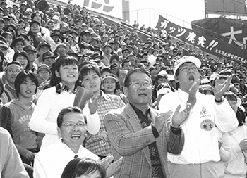 「いけるぞ、目指すは優勝だ」―。男女共学になって初の甲子園。平成８年３月２８日の第１試合、鹿児島実業高校の３塁側アルプススタンドは、初登場のチアリーダーと伝統の″野太い″応援団が見事に共演、伊都高校＝和歌山＝との接戦を選手と一緒にもぎ取った。また下窪投手の地元・指宿郡頴娃町谷場集落からは全住民の１／３が駆けつけ、「キバレ」「キバレ」の大声援。新生「カジツ」の応援と、はつらつナインが球場を圧倒した。